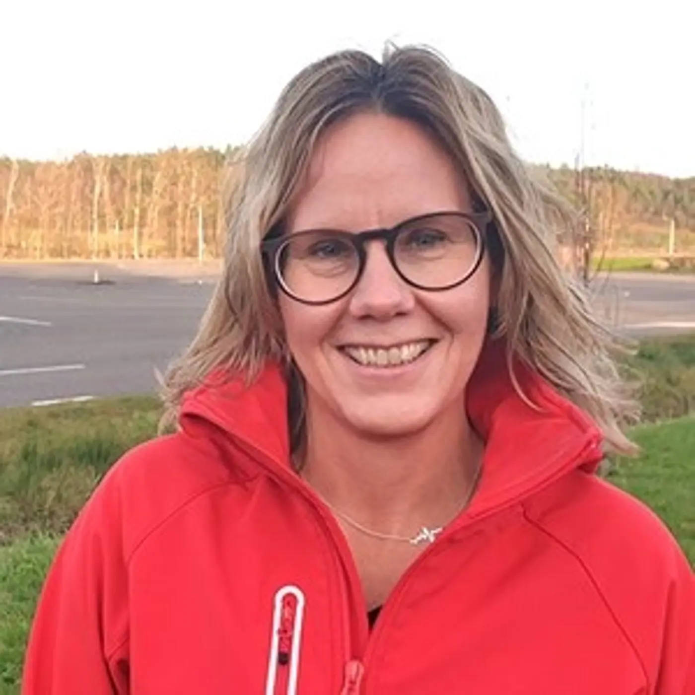 En leende kvinna med glasögon och röd jacka.