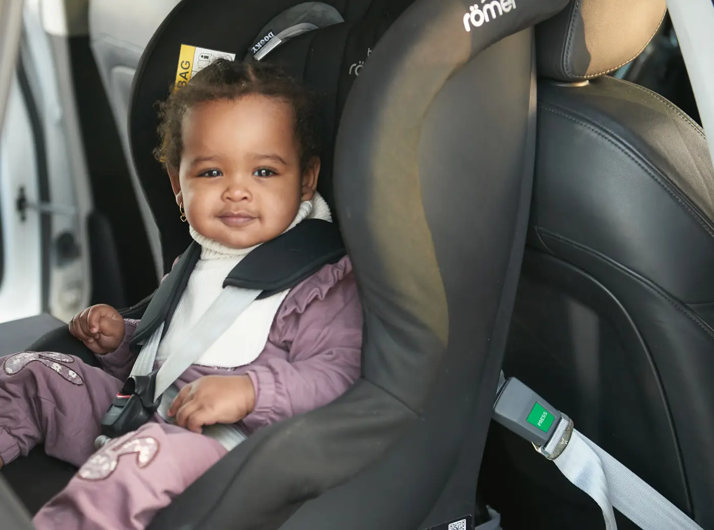 Ett småbarn sitter i en bakåtvänd bilbarnstol och tittar in i kameran.