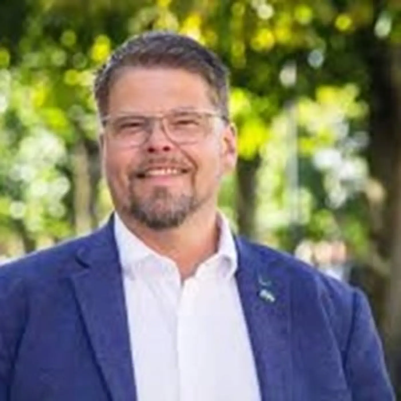 En porträttbild på en man med glasögon, vit skjorta och blå kavaj som ler mot kameran.