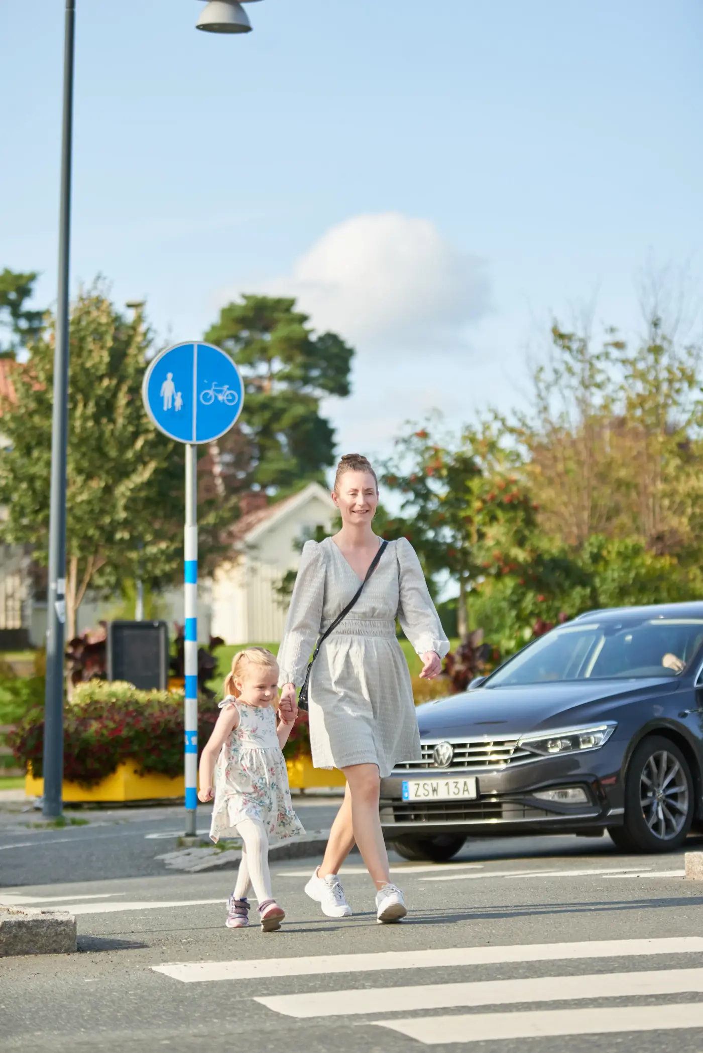 Ett barn håller sin förälder i handen och går över ett övergångsställe, framför övergångsstället väntar en bil. Bilden är tagen snett framifrån.