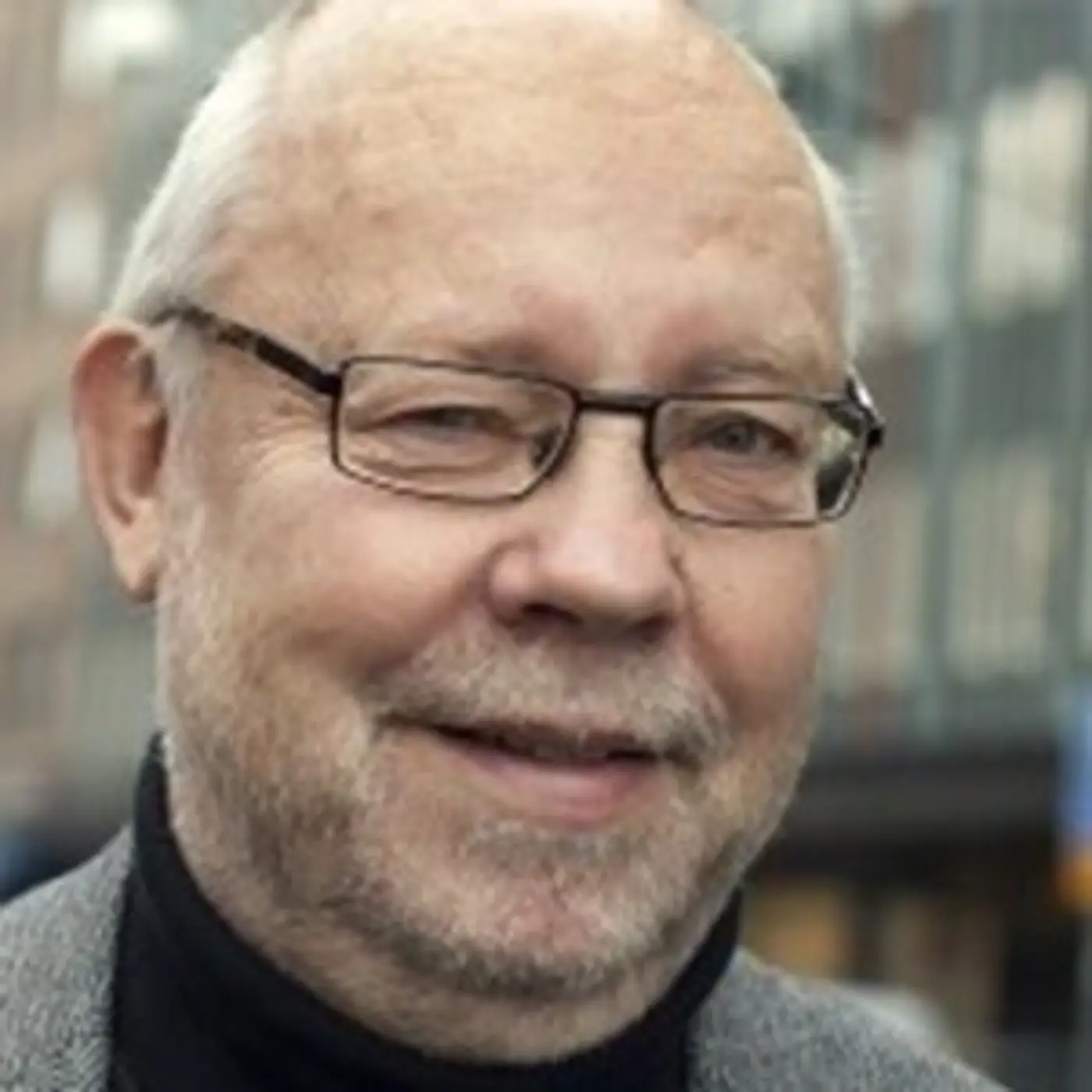 Porträttbild på en leende äldre man med glasögon och svart polotröja.