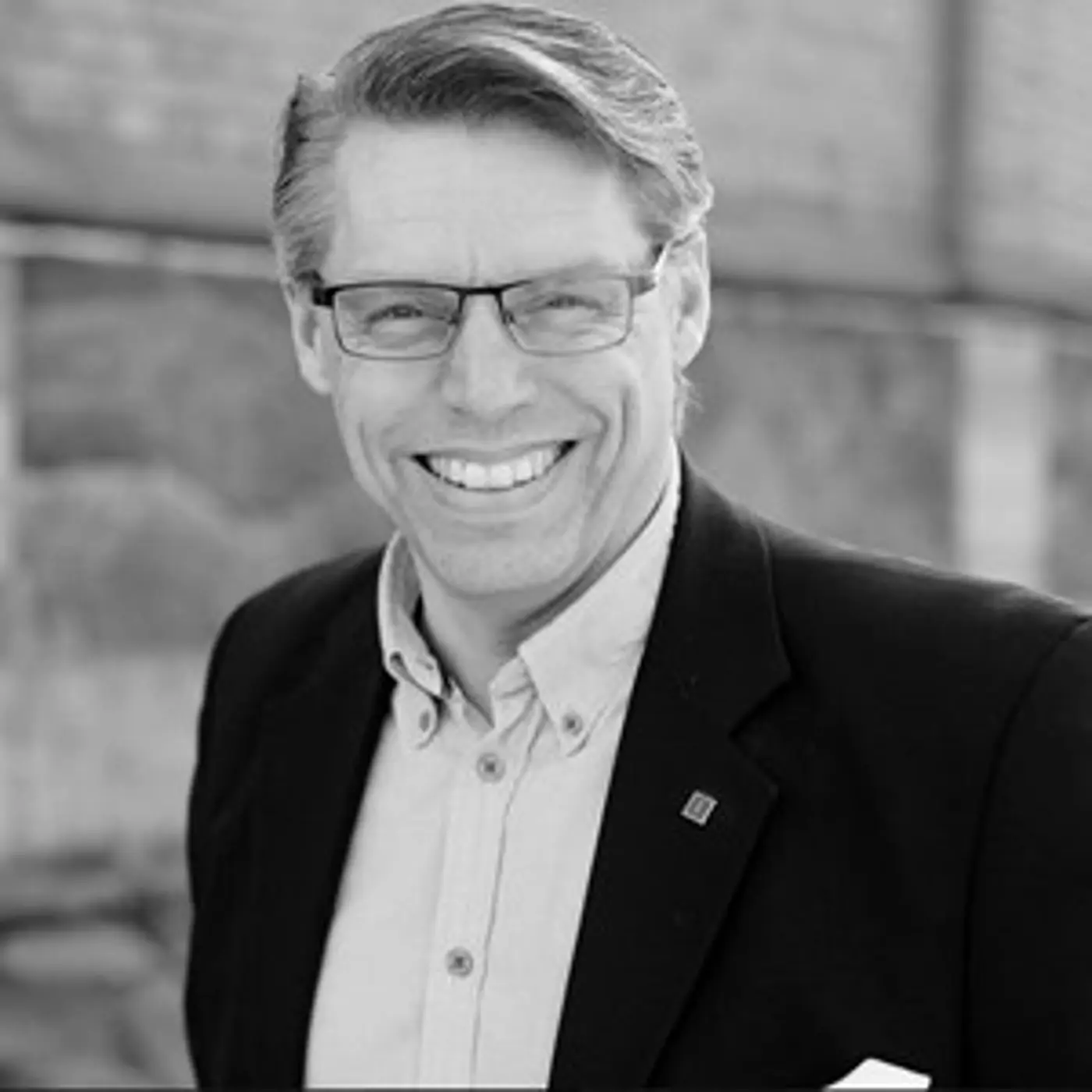 Svartvit porträttbild på en leende man med glasögon och kavaj.