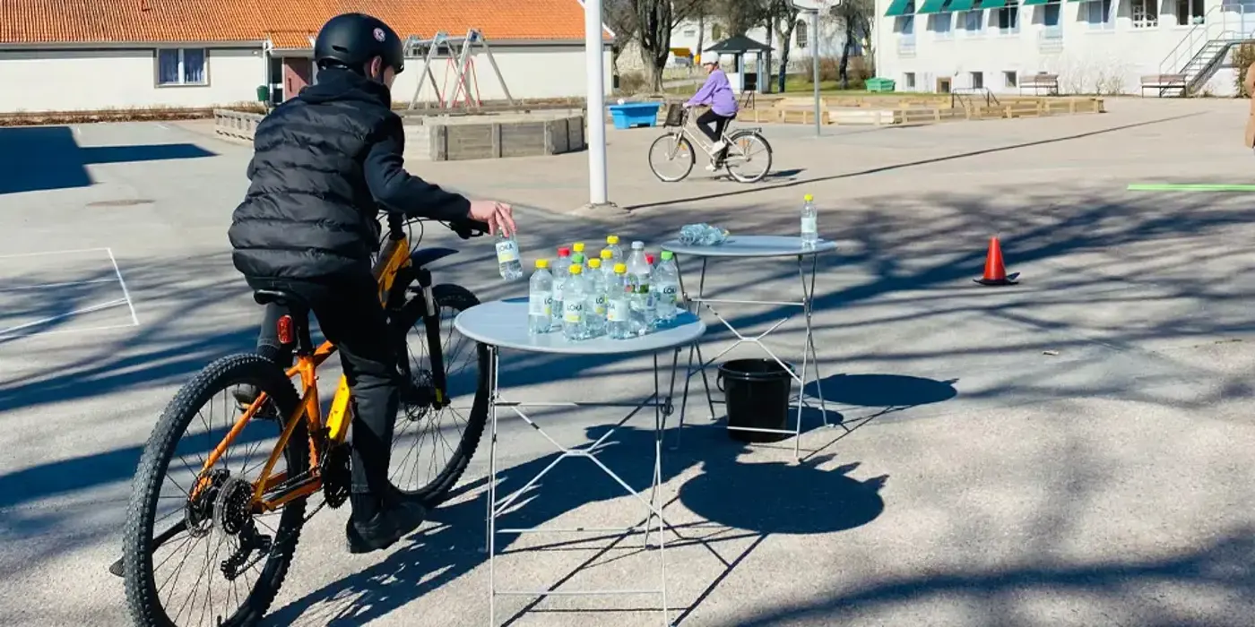 Barn kommer cyklandes på en skolgård och tar en vattenflaska som står på ett bord i farten.