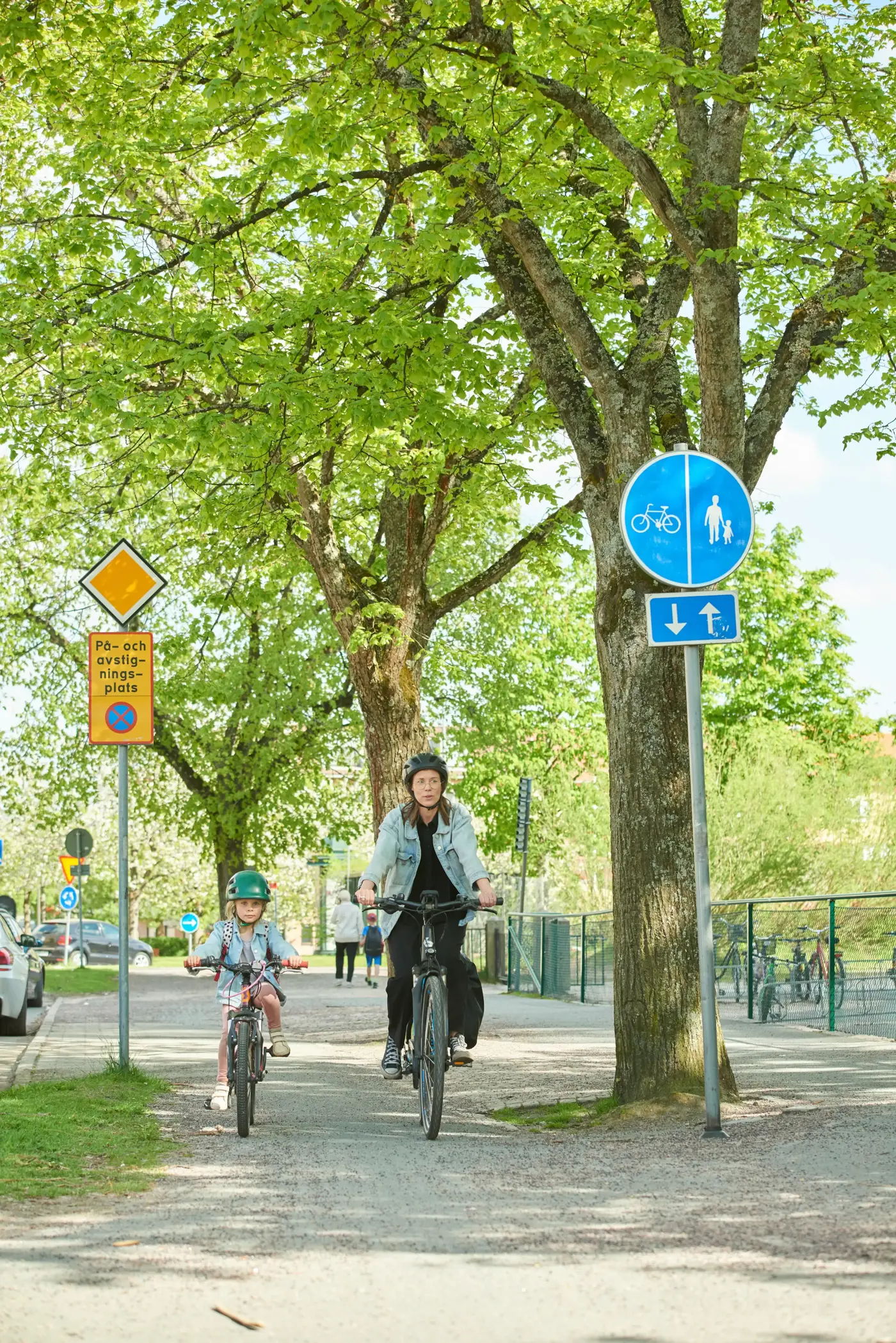 En mamma och ett barn cyklar bredvid varandra på en gång- och cykelbana utanför en skola.