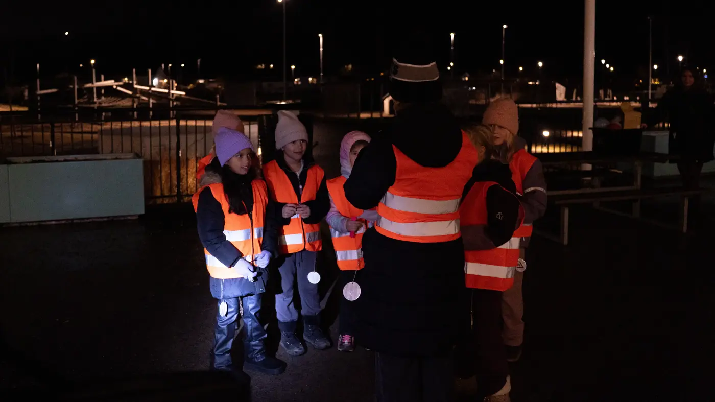 En grupp skolbarn och en lärare står utomhus i mörkret iklädda reflexvästar.