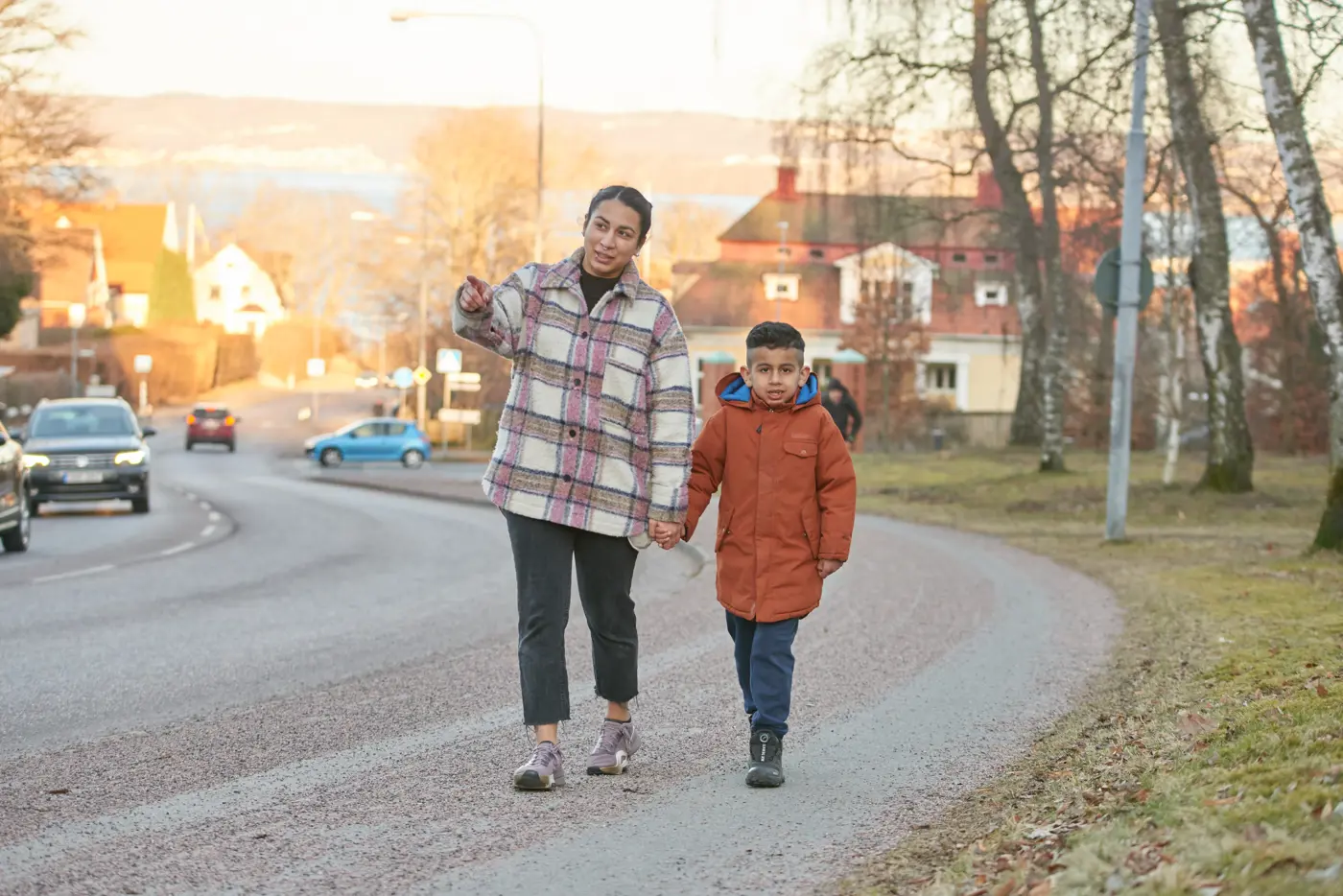 En mamma håller sitt barn i handen och går på en gångbana jämte en bilväg. Mamman går närmast vägen.