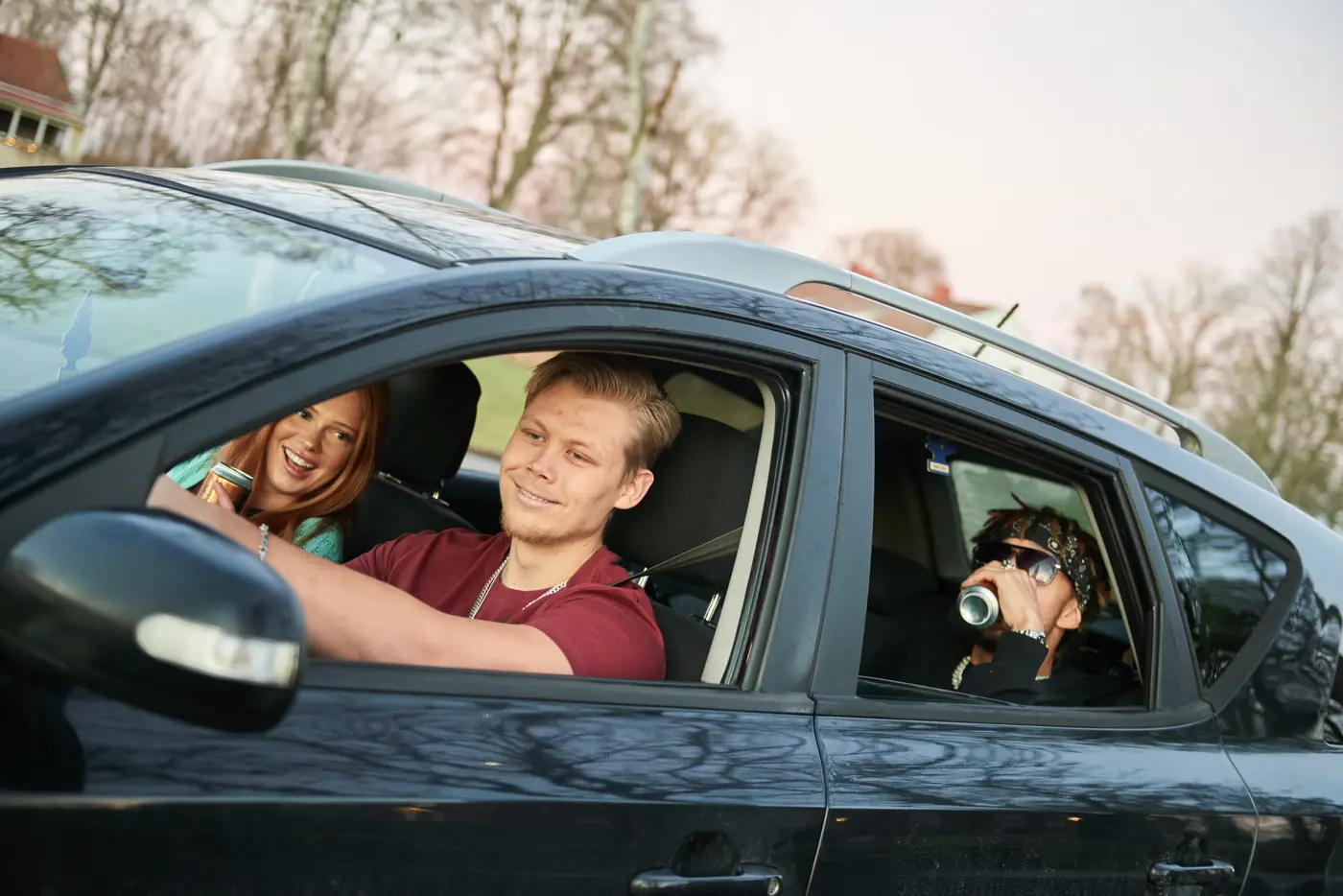 Tre ungdomar sitter i en bil. De två passagerarna dricker öl.