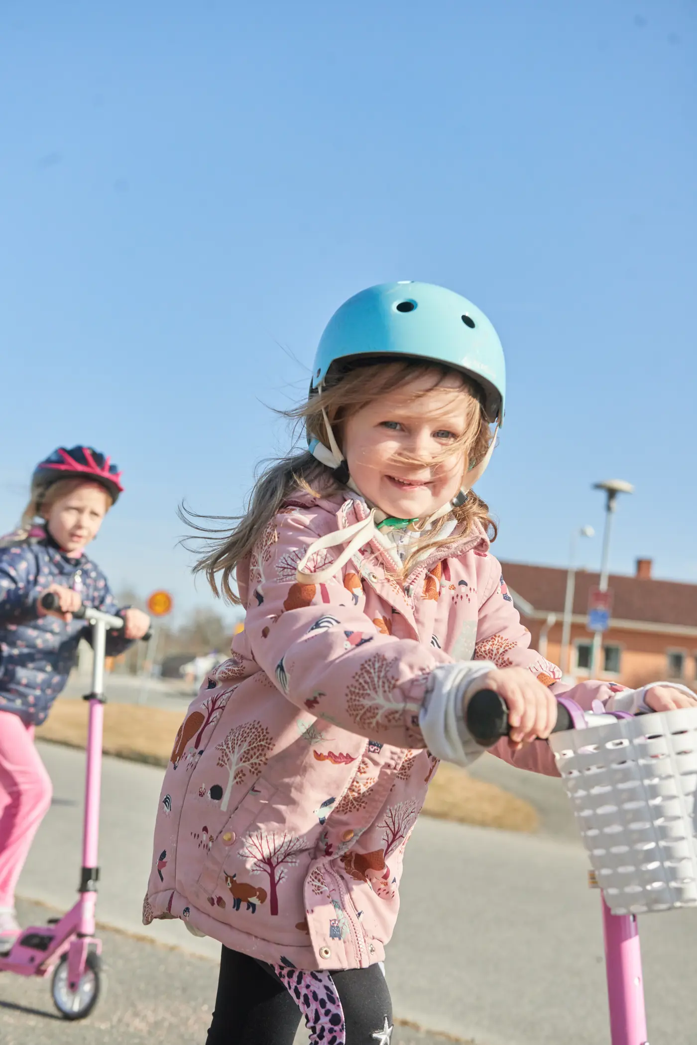 Två barn kommer cyklandes på varsin sparkcykel. Båda har hjälm på huvudet.