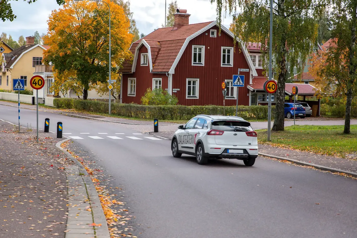 En vit bil kör på med ett villaområde till höger, närmar sig ett övergångsställe och passerar en 40-skylt.