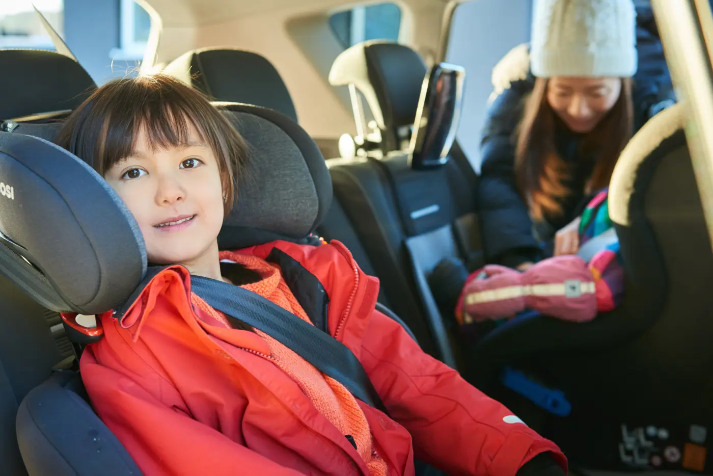 Två barn i en bil sitter i varsin bilbarnstol,  en framåtvänd och en bakåtvänd, och en mamma knäpper bältet på den ena.