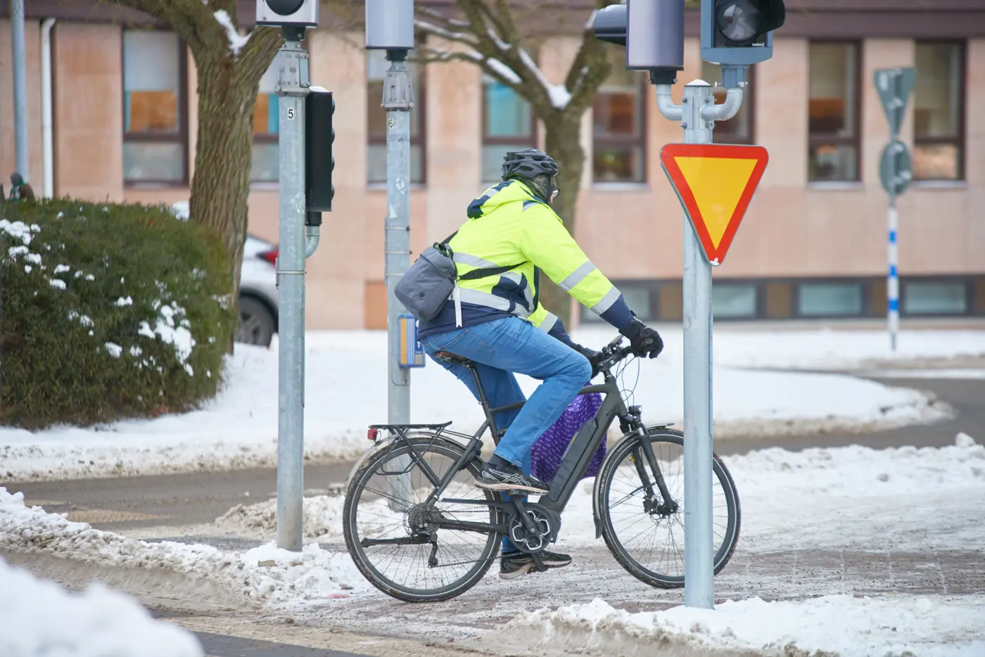 En cyklist med hjälm på huvudet och reflexjacka cyklar över ett övergångsställe i snötäckt stadsmiljö.