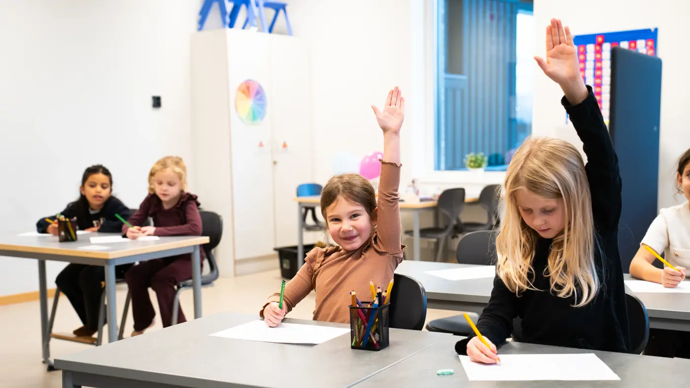 En grupp skolbarn sitter i sina bänkar och skriver och några räcker upp handen.