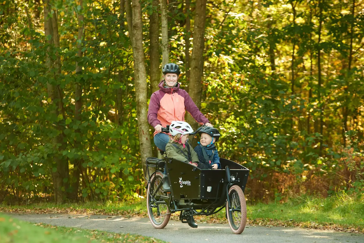 En mamma som skjutsar sina småbarn på lådcykel med cykeelhjälm och bälte.