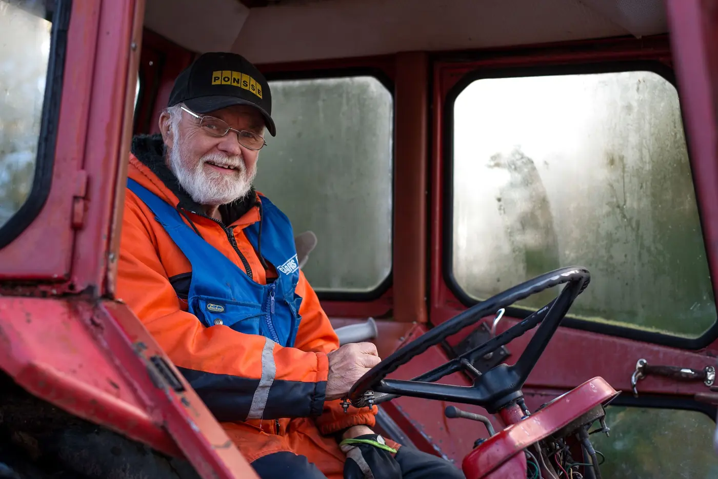 En äldre man med varselkläder sitter i en traktor och ler mot kameran