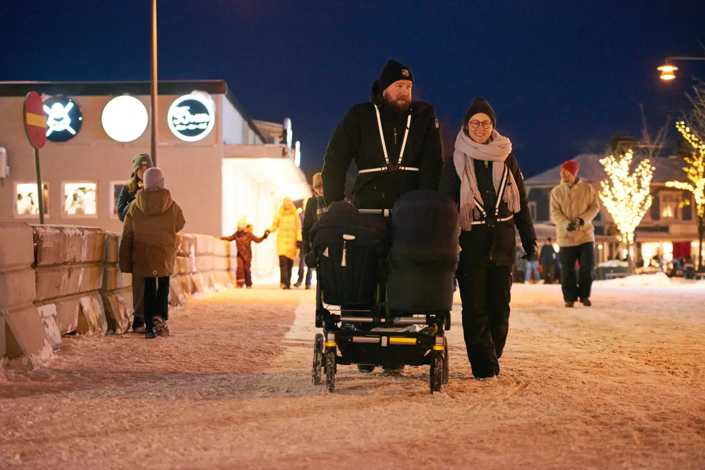 Två personer med reflexer på sig promenerar med en barnvagn på en gata i vintermiljö.