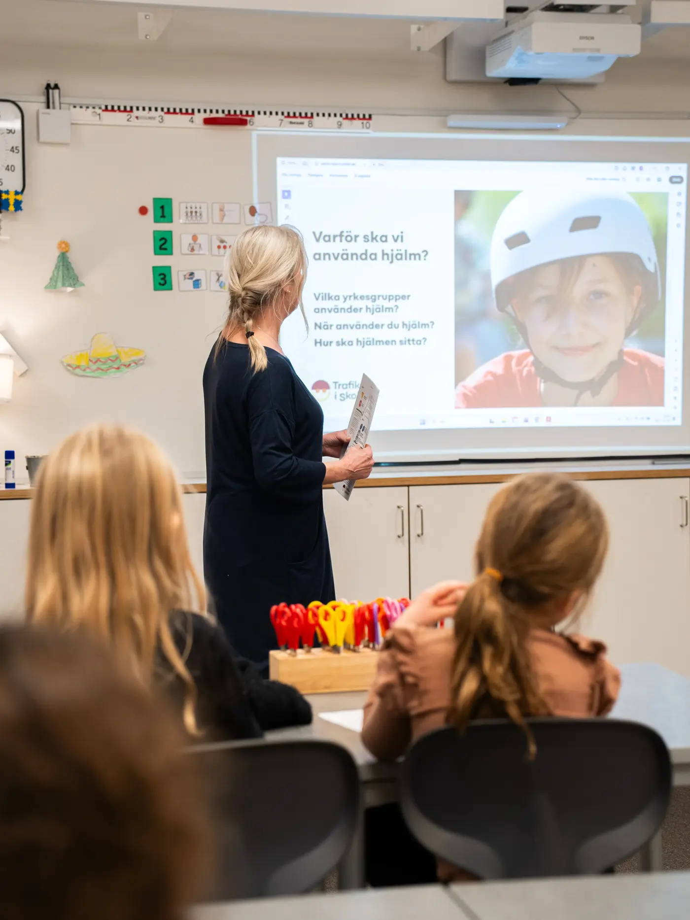 En lärare står framför en skolklass och pratar till en bild på ett barn med cykelhjälm.
