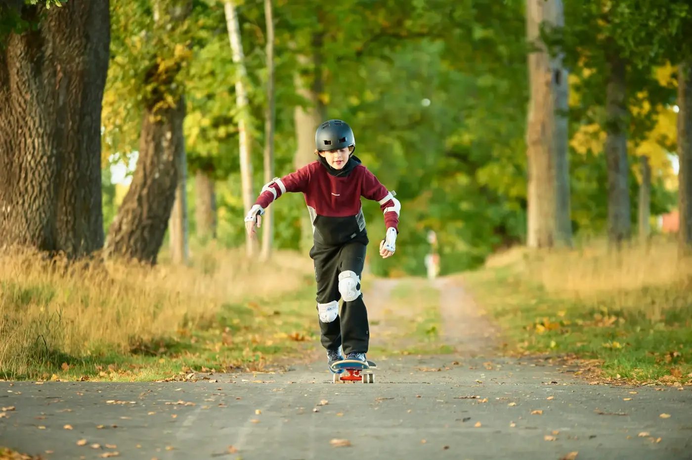 Ett barn med knäskydd, handledsskydd, armbågsskydd och hjälm kör skateboard på en väg omgiven av en allé med träd.
