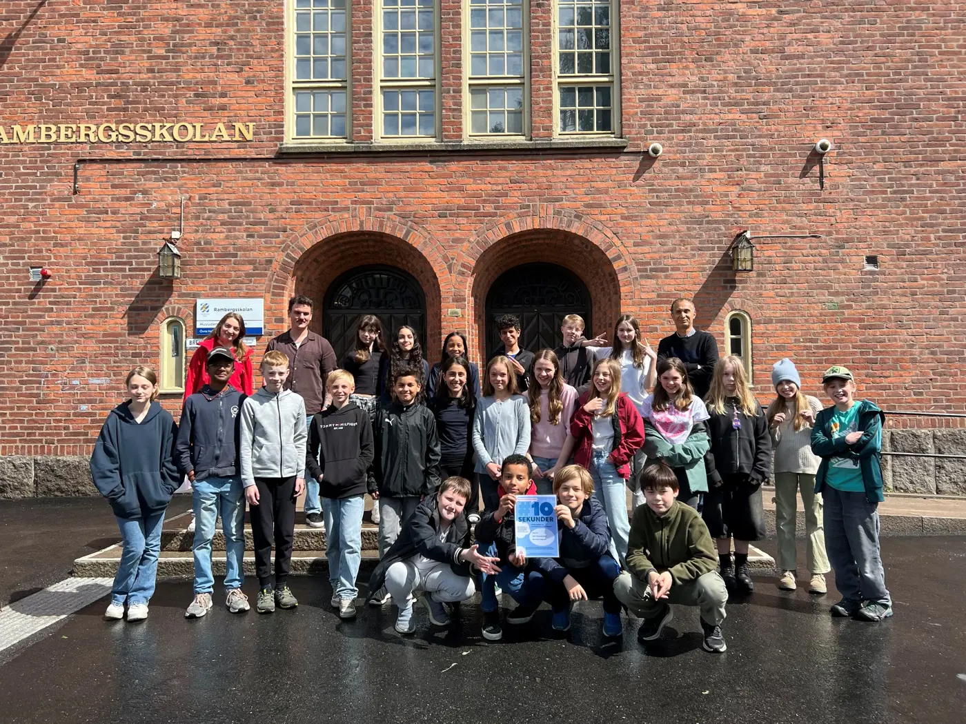 Skolklass står framför ingången till Rambergsskolan och håller upp ett diplom.