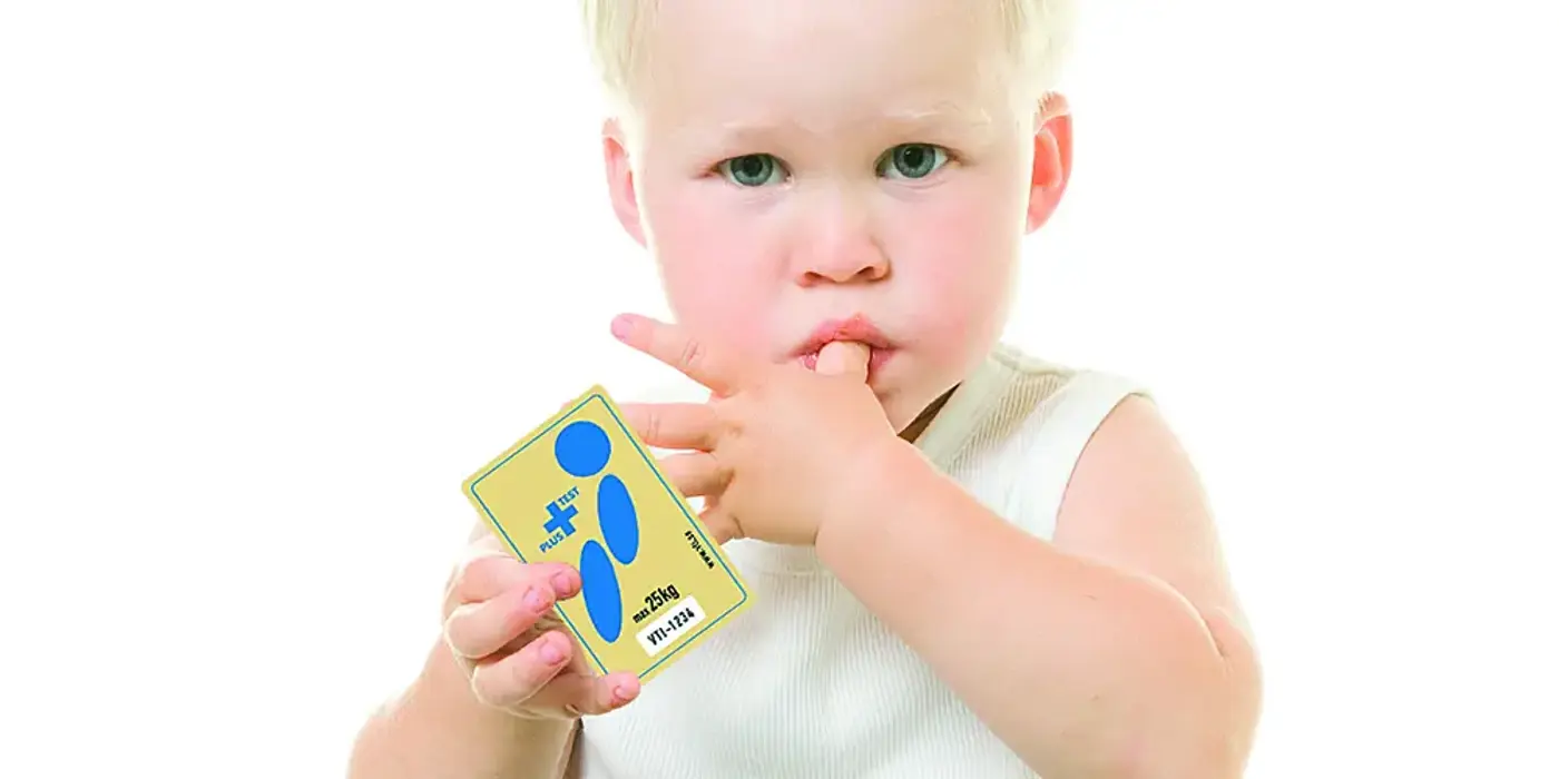 Ett litet barn tittar in i kameran, suger på tummen och håller i ett kort.