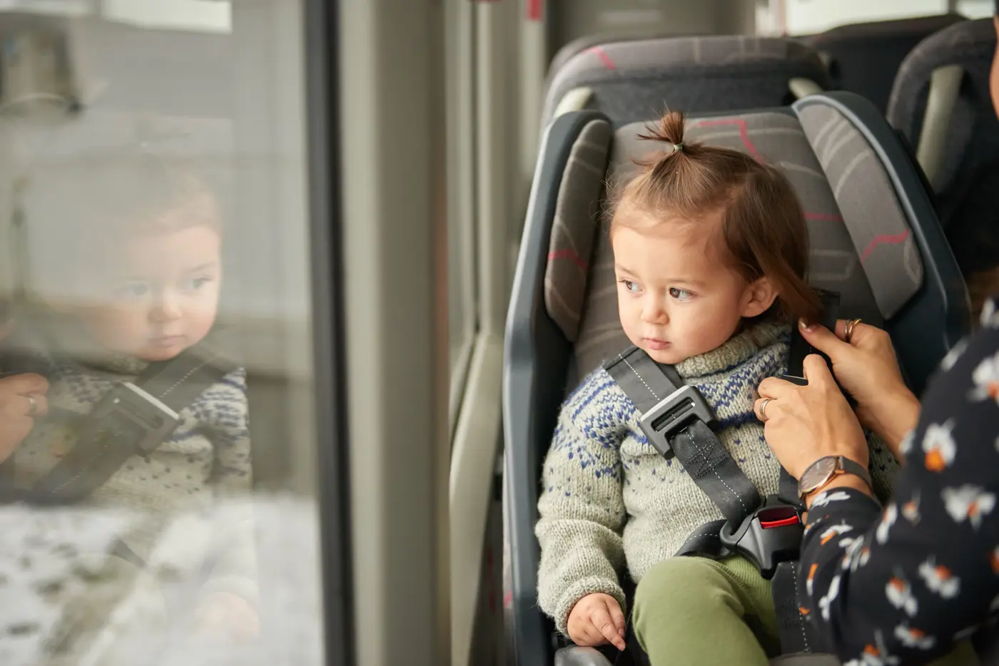 Ett småbarn sitter i en bakåtvänd bilbarnstol i en buss samtidigt som en förälder håller på att spänna bältet.