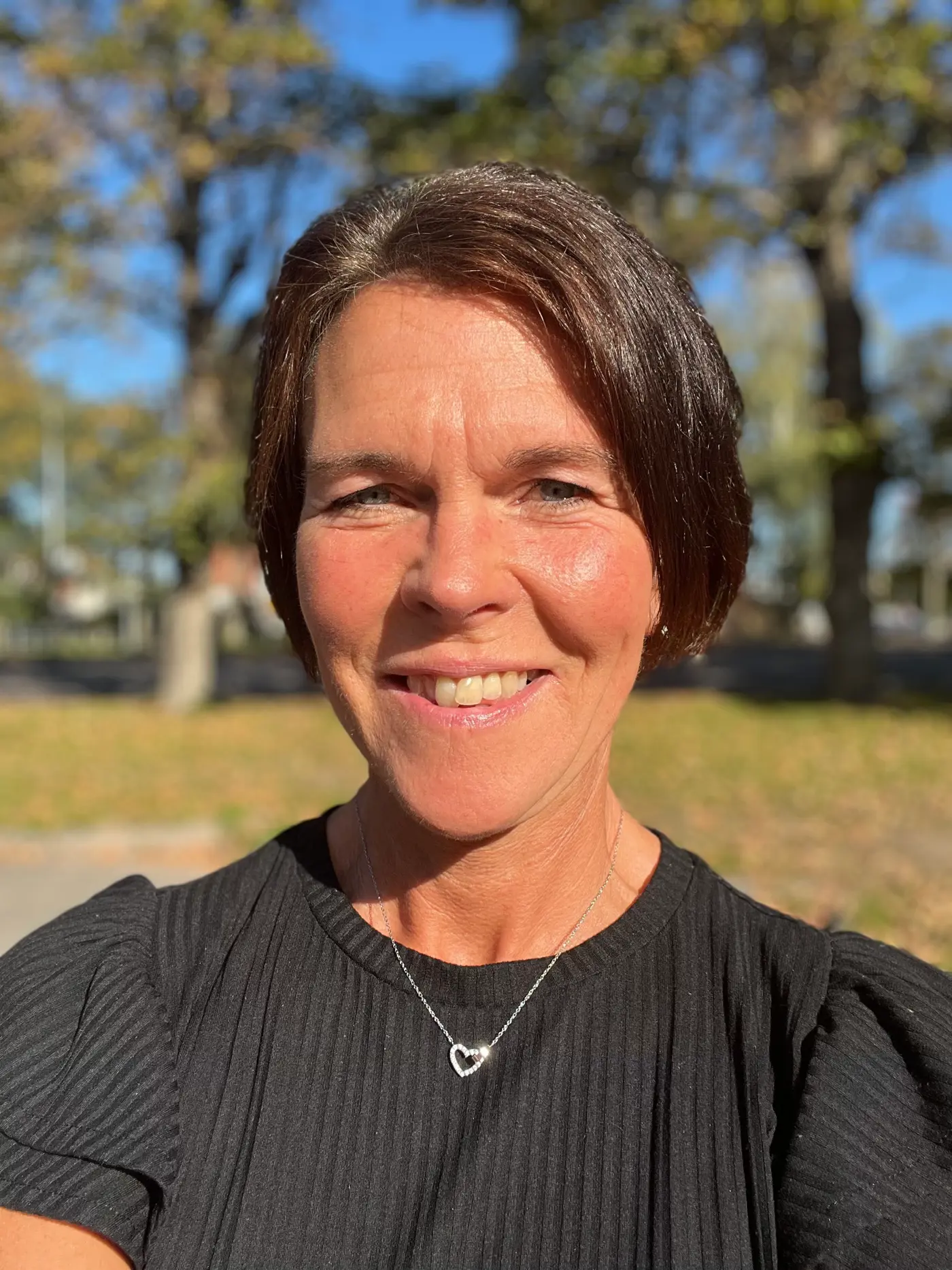 En porträttbild på en leende kvinna med kort hår och svart tröja. Hon står utomhus i solljus.