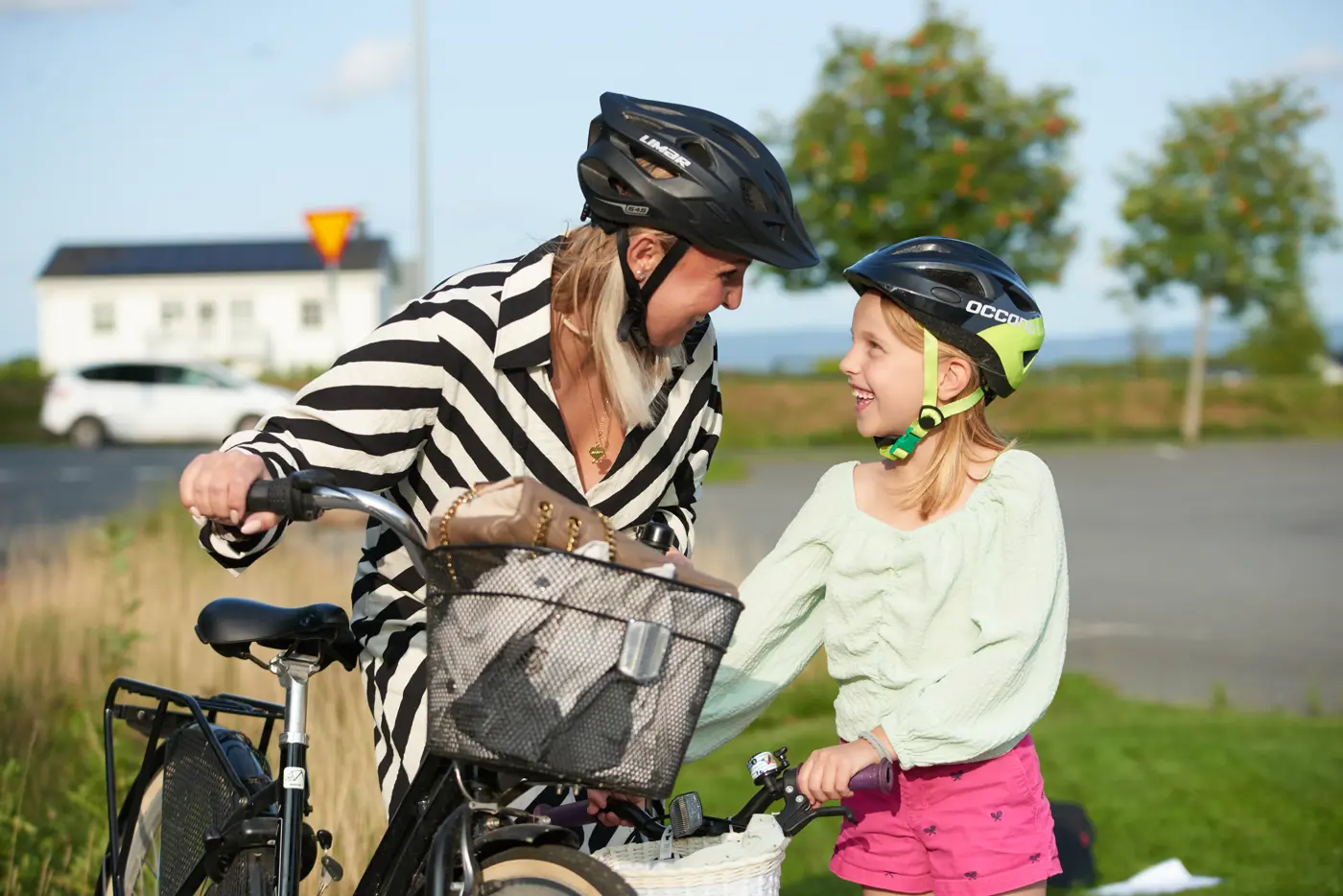 En mamma och dotter står med varsin cykel, tittar på varandra och skrattar. De har båda hjälm på huvudet.