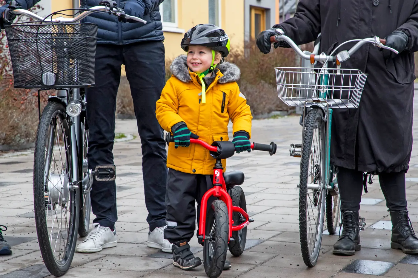 Ett barn med hjälm på huvudet leder en balanscykel. Barnet går mellan två vuxna som även de leder varsin cykel.