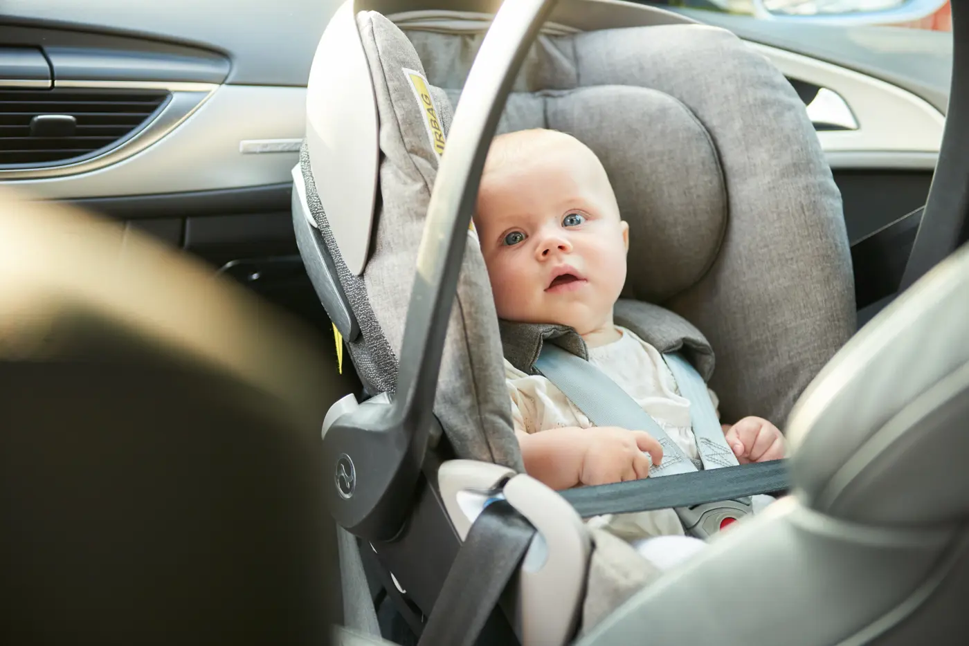 En bebis sitter i ett babyskydd i framsätet på en bil och tittar mot kameran.