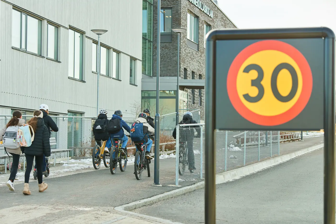 Åtta skolbarn är på väg mot en skola, fem cyklar och tre promenerar. I förgrunden syns en 30-skylt.