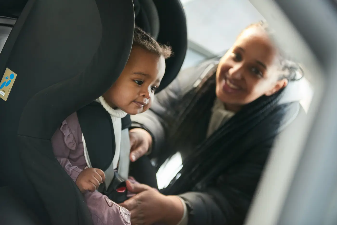 Ett småbarn sitter i en bakåtvänd bilbarnstol och barnets mamma håller på att knäppa fast bältet.