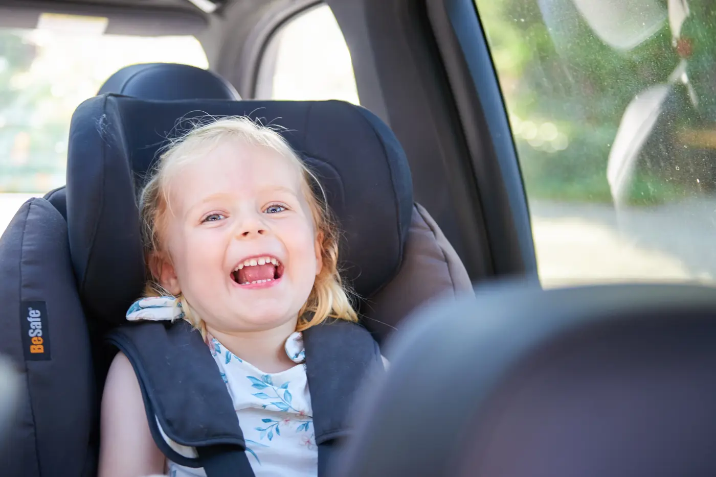 Ett barn sitter i en bakåtvänd bilbarnstol och skrattar mot kameran.