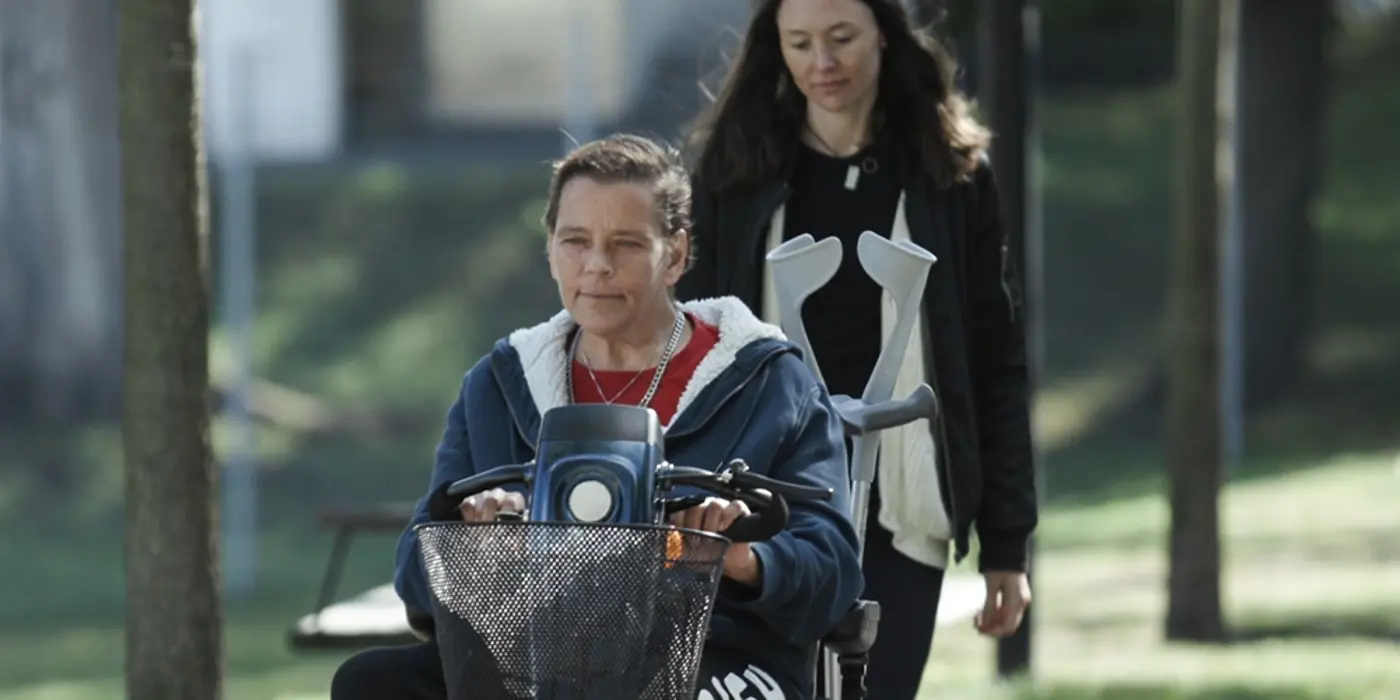 En person kör en elskoter på en väg i ett grönområde. Bakom promenerar ytterligare en person.