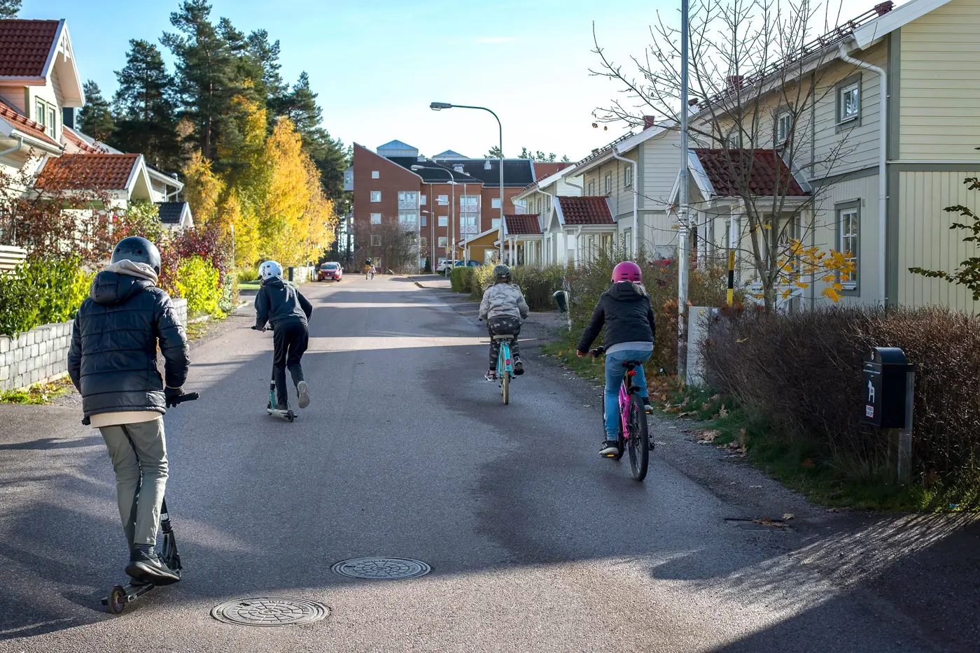 Fyra barn kör varsitt fordon på en asfalterad väg i ett bostadsområde, två av barnen cyklar och två kör kickbike. Alla har hjälm på huvudet.